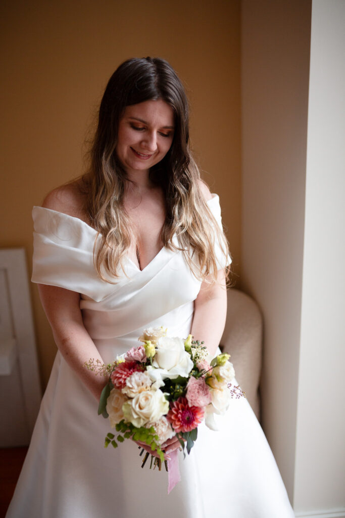 mariée portant son bouquet devant une fenêtre. la chambre est éclairée avec la lumière natrelle du jour