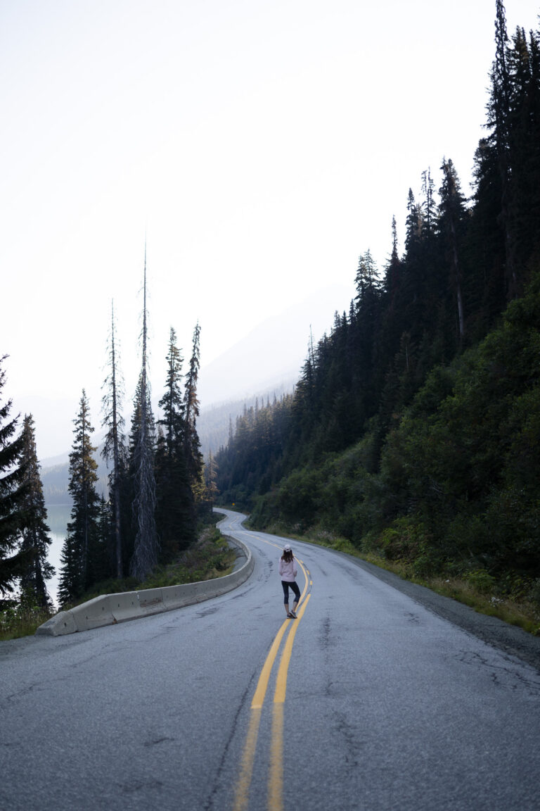 Personne marchant au centre d’une route de montagne bordée d’une forêt de conifères et d’un lac, dans un paysage brumeux de l’Alberta au Canada.